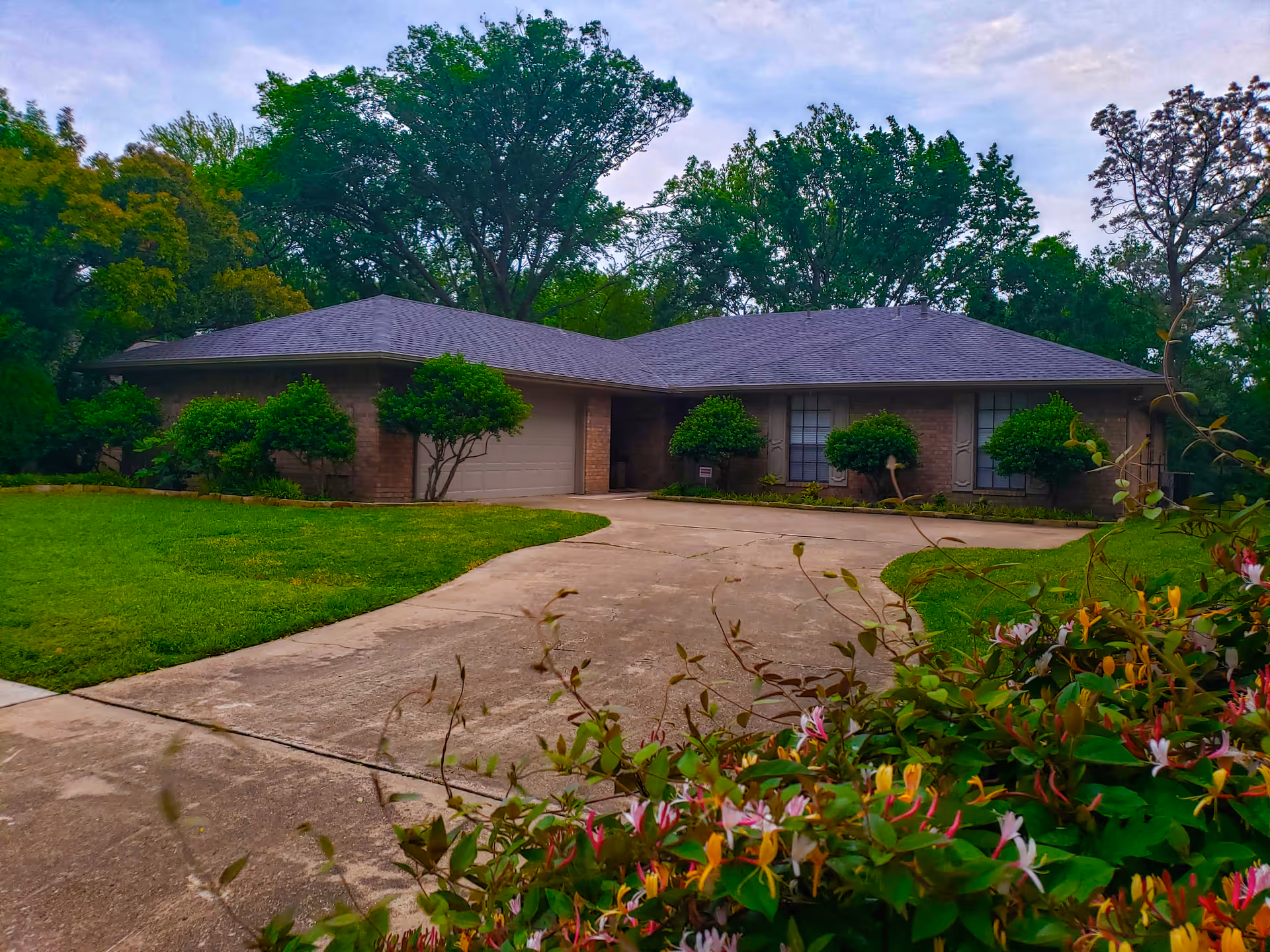A single-story brick building with a gray roof surrounded by green trees and bushes. There is a curved concrete driveway leading to a garage on the left side of the building. Flowering plants are visible in the foreground.