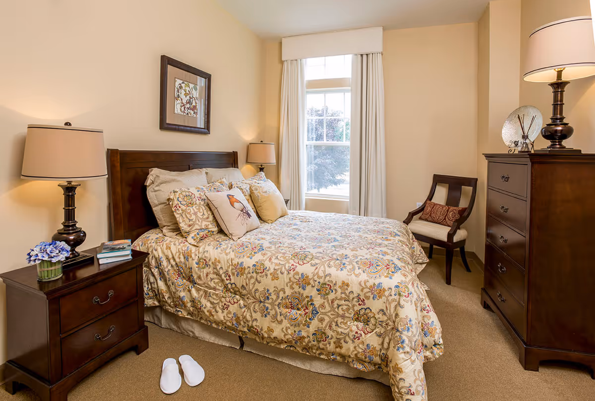 A cozy bedroom with a wooden bed frame and floral patterned bedding. There are multiple decorative pillows on the bed, including one with a bird design. On the left side, there is a wooden nightstand with a lamp, a small plant with purple flowers, and some books. White slippers are placed on the beige carpet near the bed. A window with white curtains allows natural light into the room. On the right side, there is a wooden dresser with a lamp and decorative items on top, and a wooden chair with a cushion and a decorative pillow.