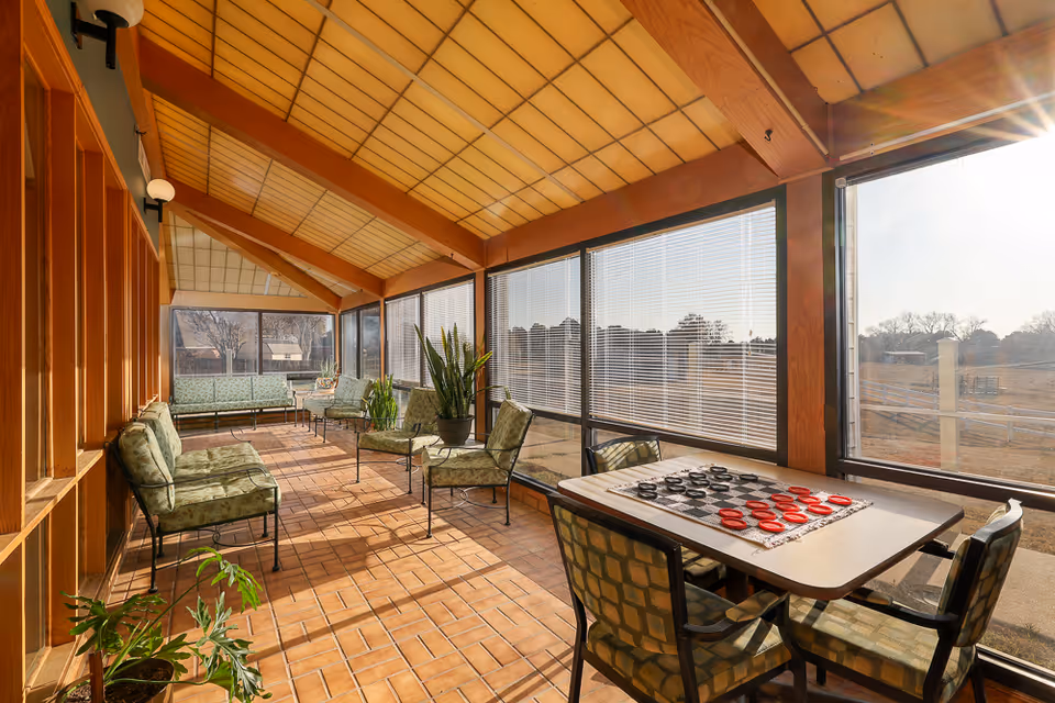 A sunlit enclosed porch or sunroom with large windows and blinds, featuring several cushioned chairs and sofas arranged along the walls, a table with a checkers game set up, and potted plants placed around the space. The ceiling has wooden beams and a warm-toned panel design, and the floor is tiled with terracotta tiles.