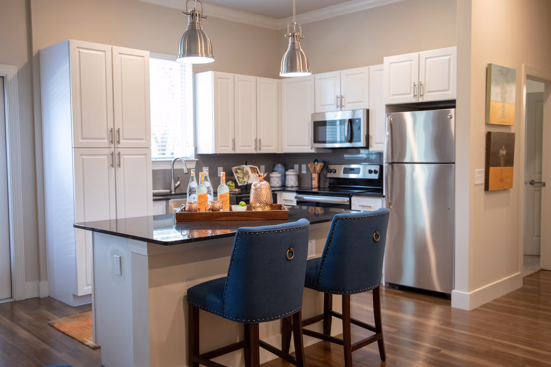 Modern kitchen with white cabinets, stainless steel refrigerator and microwave, black countertop island with two blue upholstered bar stools, and pendant lights hanging above the island. A tray with bottles and decorative items is placed on the island. Hardwood flooring and neutral wall colors complete the space.