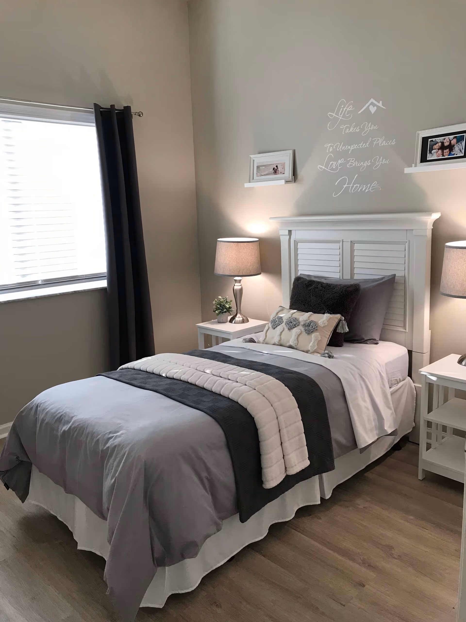 A neatly made single bed with gray and white bedding in a bedroom. The bed has multiple pillows and a white headboard. There are two white nightstands on either side of the bed, each with a lamp and one with a small plant. A window with dark curtains is on the left wall. Above the bed, there is a wall decal with the words: 'Life Takes You To Unexpected Places Love Brings You Home.' Two small shelves with framed photos are mounted on the wall above the headboard.
