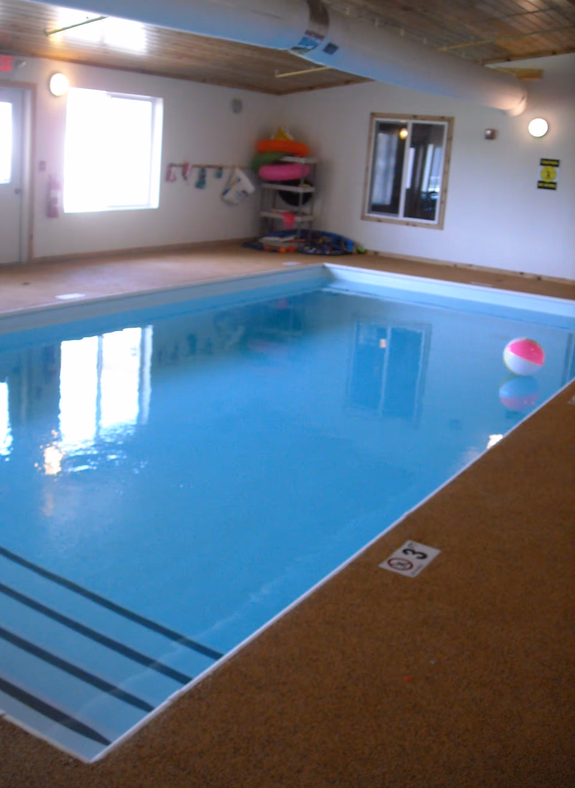 Indoor swimming pool with clear blue water, a beach ball floating on the surface, steps leading into the pool, and pool safety signs on the wall. The room has a wooden ceiling, a window, and a door letting in natural light. Pool toys and floatation devices are stacked on shelves in the corner.