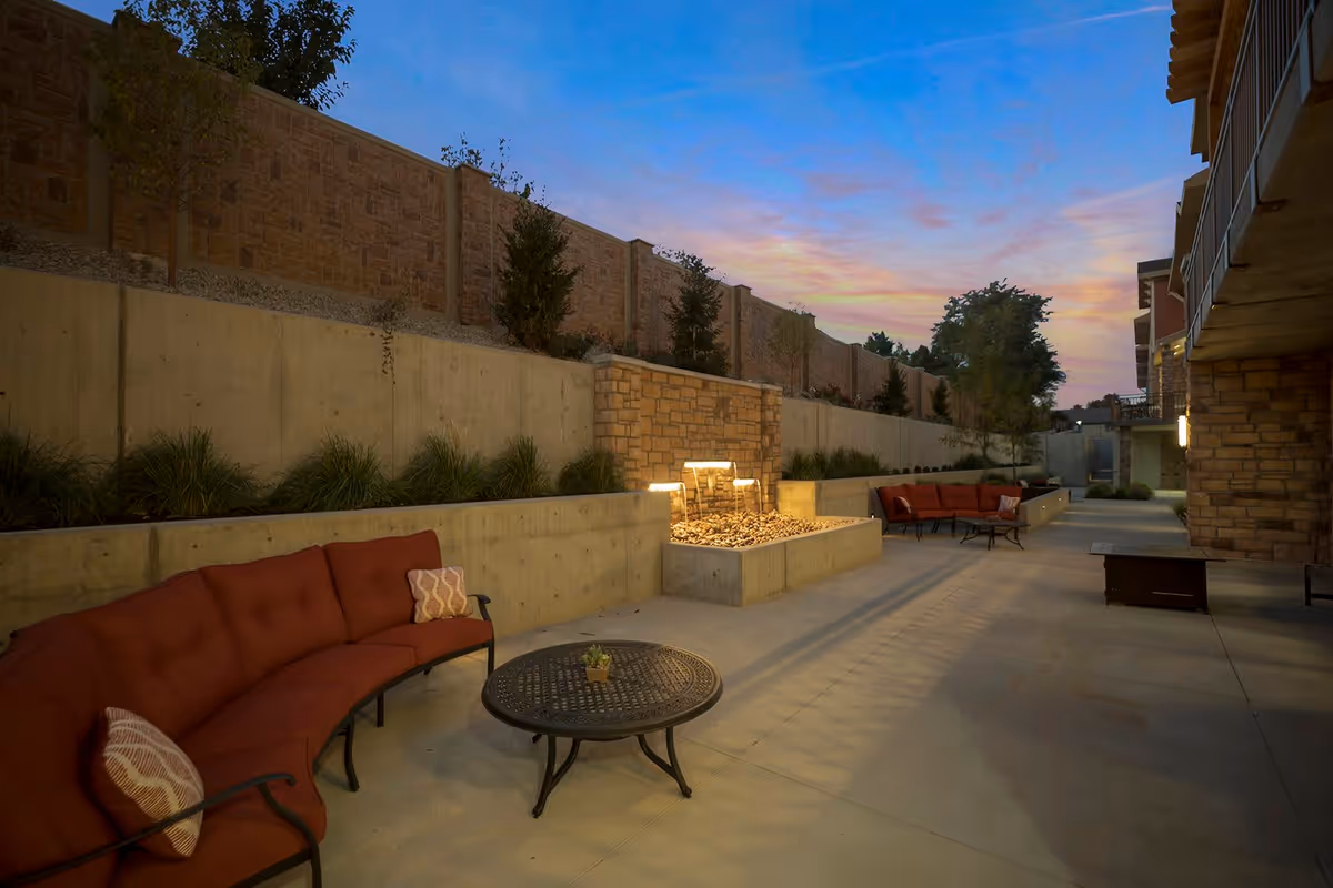Outdoor patio area at dusk with curved red cushioned seating, a round metal table with a small plant, a stone water feature with lights, and a concrete wall with landscaping and trees.