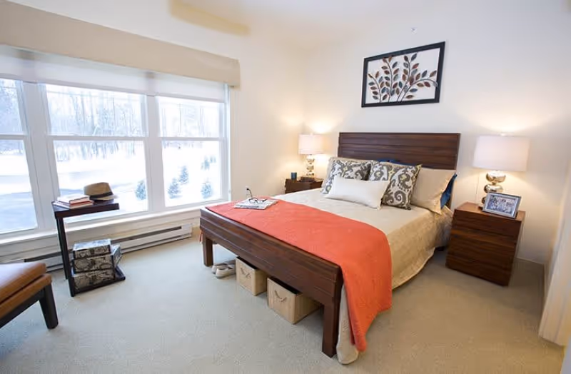 A bright bedroom with a large window showing a snowy outdoor scene. The room features a wooden bed with beige bedding and a coral throw blanket, two matching wooden nightstands with lamps, and decorative pillows. There is a framed artwork above the bed and a small table with books and a hat near the window.