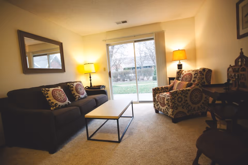 A cozy living room with a dark sofa adorned with patterned cushions, a matching patterned armchair, a wooden coffee table, two lit table lamps, a large mirror on the wall, and a sliding glass door leading to an outdoor area with trees and bushes.