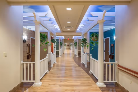 A brightly lit indoor hallway with wooden flooring and white columns supporting a pergola-style ceiling with a sky mural. Hanging plants are suspended from the pergola, and the hallway is flanked by walls painted in different colors with doors and decorative elements.