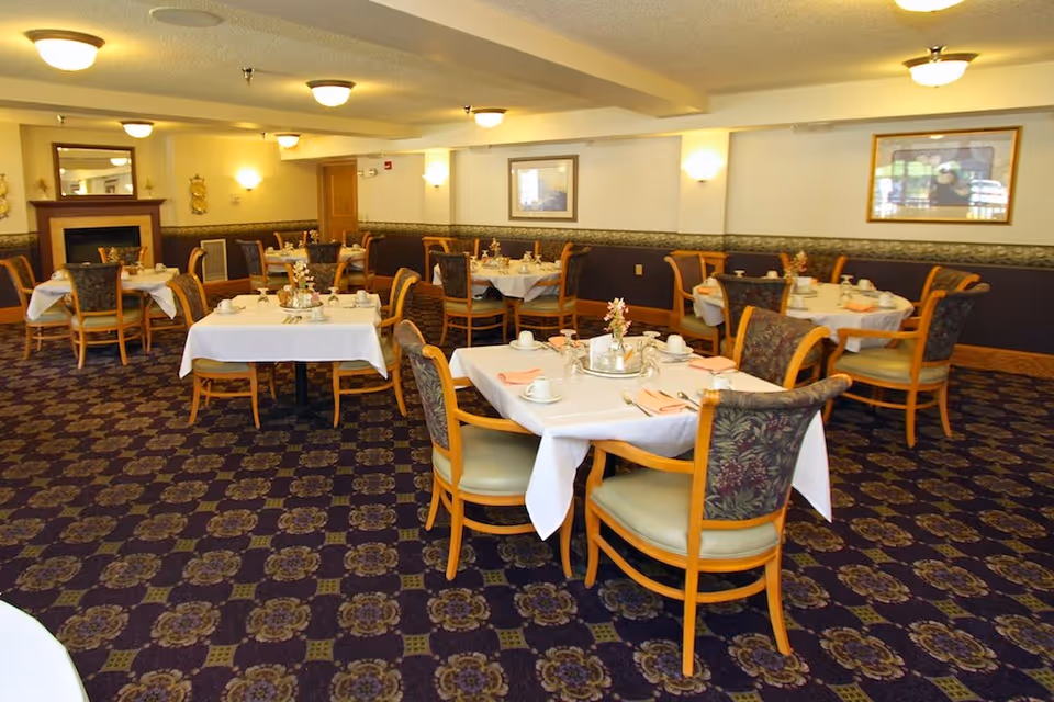 Dining room with multiple tables set for meals, wooden chairs, floral centerpieces and patterned carpet.