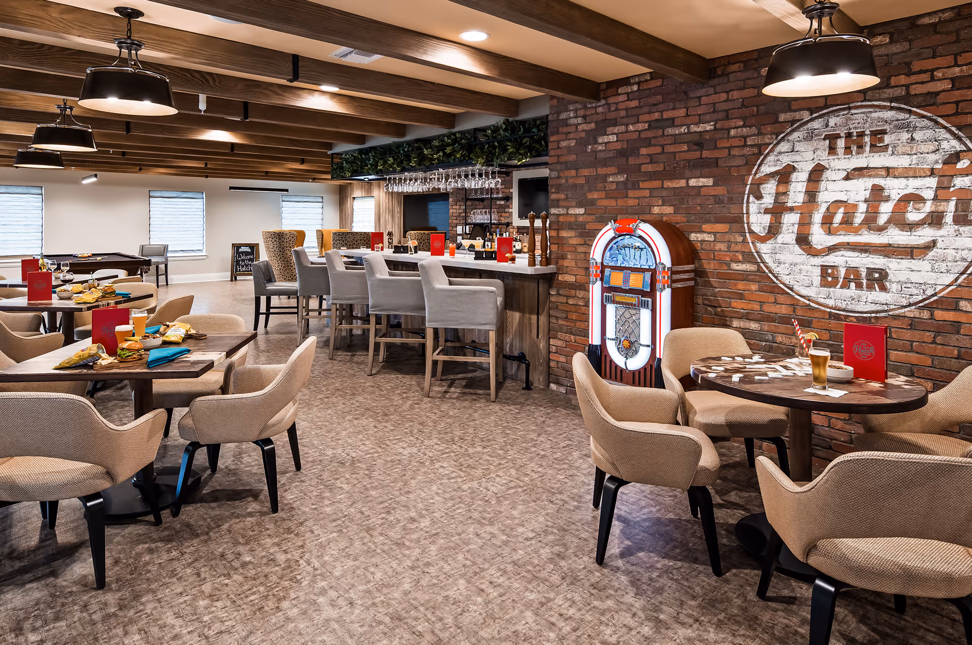 Cozy dining and bar area with tables and chairs, a central bar, a jukebox, and a painted brick sign reading 'The Hatch Bar'.