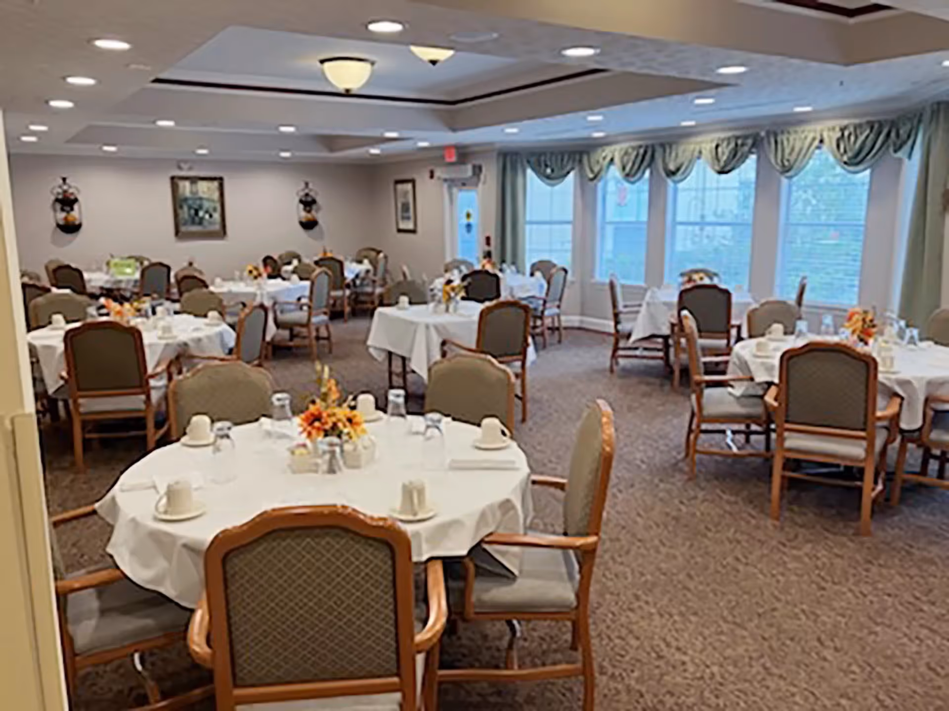 Communal dining room with round tables covered in white tablecloths, chairs, and floral centerpieces near large windows.