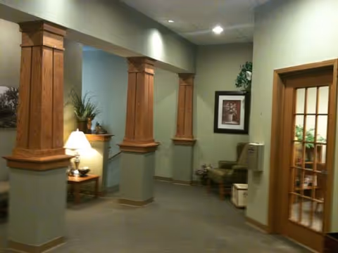 Interior hallway with green walls and wooden columns featuring decorative trim. There is a small table with a lamp and plant on the left side, a framed picture on the wall, and a seating area with a chair and small table near a water dispenser. A wooden door with glass panels is visible on the right.