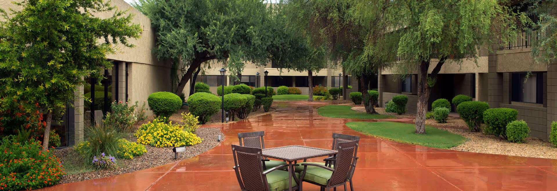 Outdoor courtyard area with a wet red concrete pathway, surrounded by green bushes, trees, and colorful flowers. There is a table with four chairs featuring green cushions in the foreground. The courtyard is enclosed by beige buildings with windows.