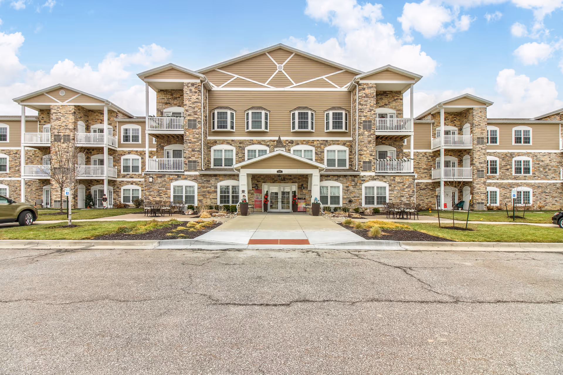 Front exterior view of a three-story senior living facility with stone and beige siding, multiple balconies, and a covered entrance with a walkway leading to the parking area under a partly cloudy sky.