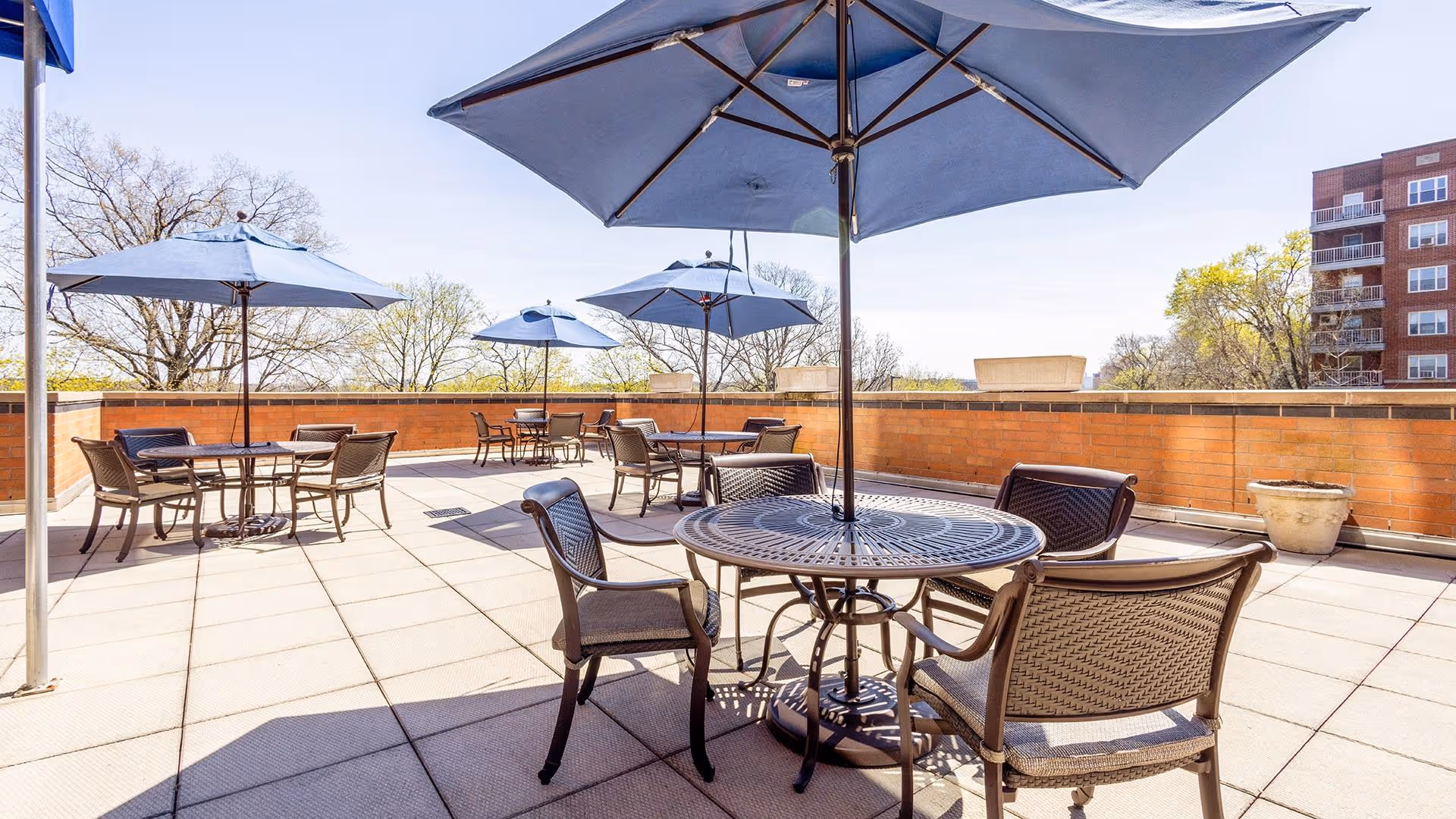 Outdoor patio area with multiple round metal tables and chairs, each shaded by large blue umbrellas. The patio is surrounded by a low brick wall, with trees and a multi-story building visible in the background under a clear sky.