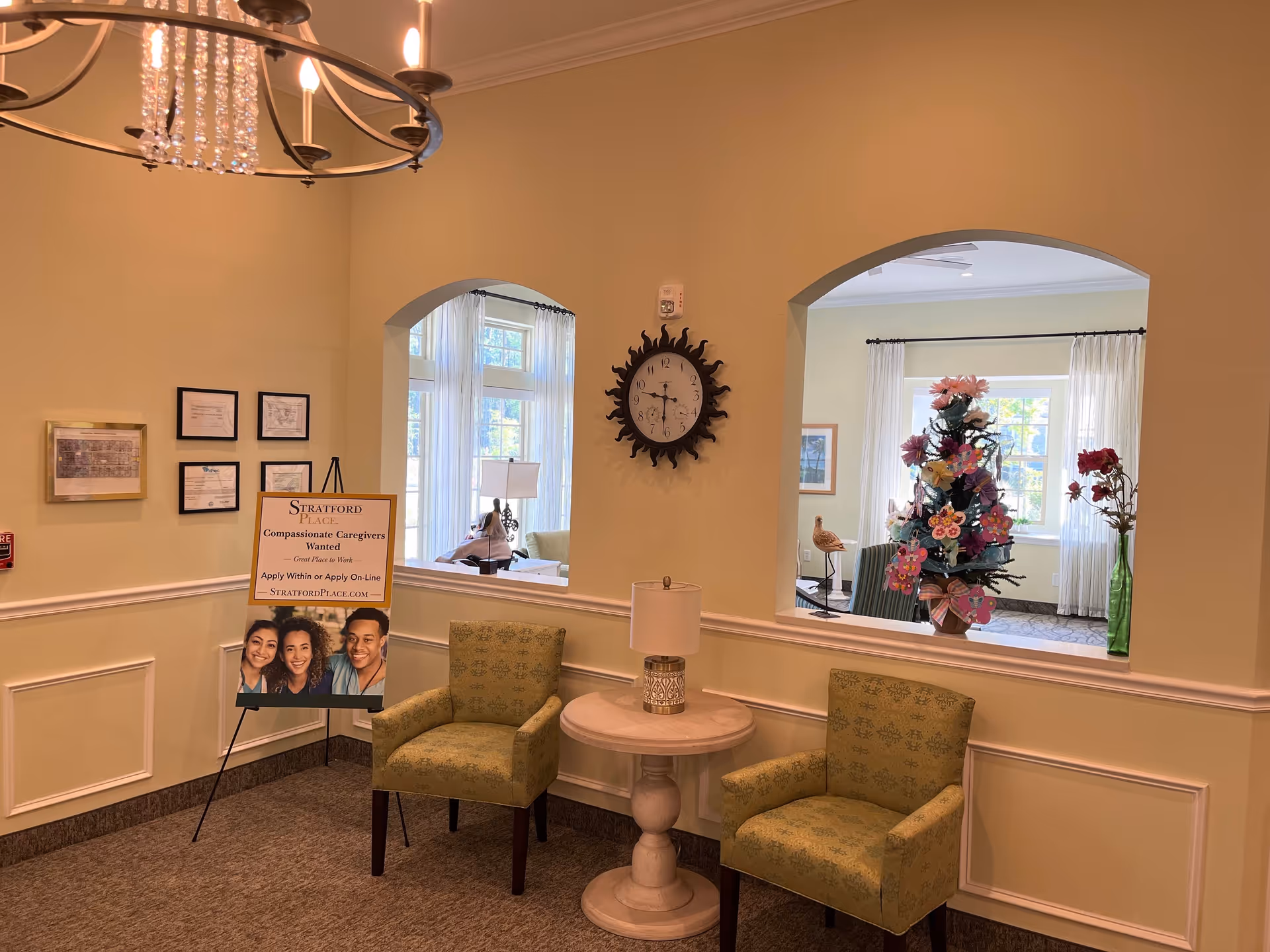 A cozy seating area in an assisted living facility with two green patterned armchairs and a round wooden table with a lamp between them. On the wall above the table is a decorative clock with a sunburst design. Two arched windows in the wall provide a view into another room with large windows, sheer white curtains, a decorated Christmas tree, and a person sitting on a couch. A sign on an easel reads 'Stratford Place Compassionate Caregivers Wanted - Great Place to Work - Apply Within or Apply On-Line - StratfordPlace.com'. Several framed certificates and documents are hung on the wall to the left.