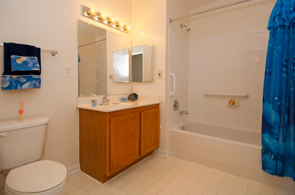 A clean bathroom featuring a white toilet, a wooden vanity with a sink, a large mirror with lights above it, and a bathtub with a blue shower curtain. The walls and floor are light-colored, and there are two towels hanging on a rack above the toilet.