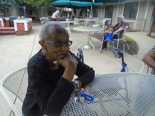 An elderly woman wearing glasses and a black top sits at a metal outdoor table with her hand resting on her chin. In the background, other elderly individuals are seated or standing near similar tables and chairs on a paved patio area outside a building.