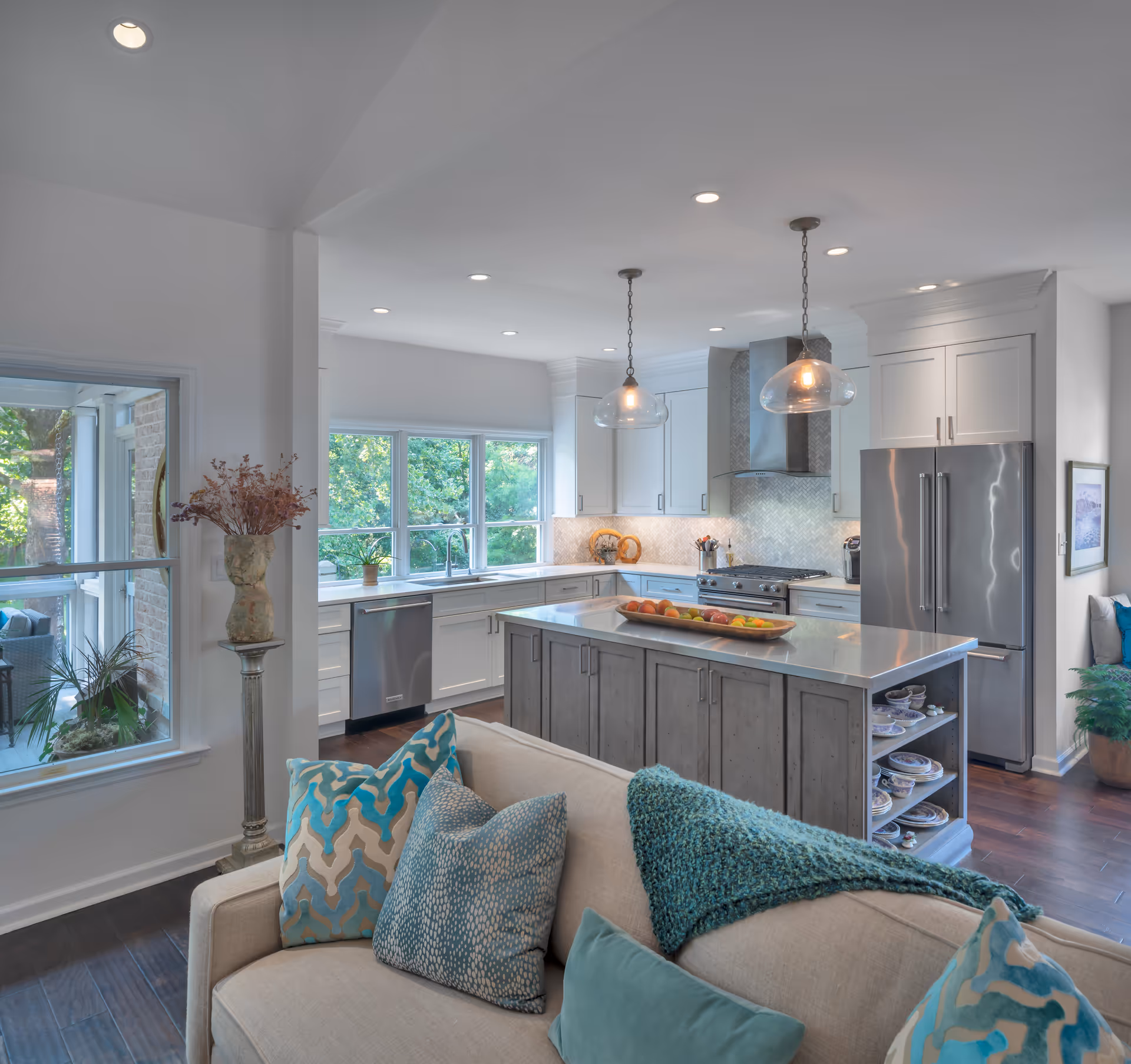 Bright and modern kitchen with white cabinetry, stainless steel appliances including a refrigerator and dishwasher, a large island with a fruit tray, pendant lights hanging above, and a beige sofa with blue and teal pillows in the foreground. Large windows provide a view of green trees outside.