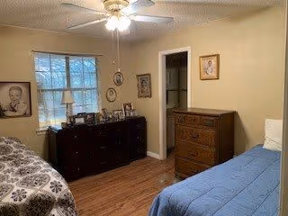 A bedroom with two beds, one with a blue bedspread and the other with a patterned bedspread. There is a ceiling fan with a light fixture above. A window with blinds is on the left wall, and below it is a dark wooden dresser with framed photos and a lamp. The walls are decorated with several framed pictures. There is a wooden chest of drawers against the right wall and a doorway leading to another room.