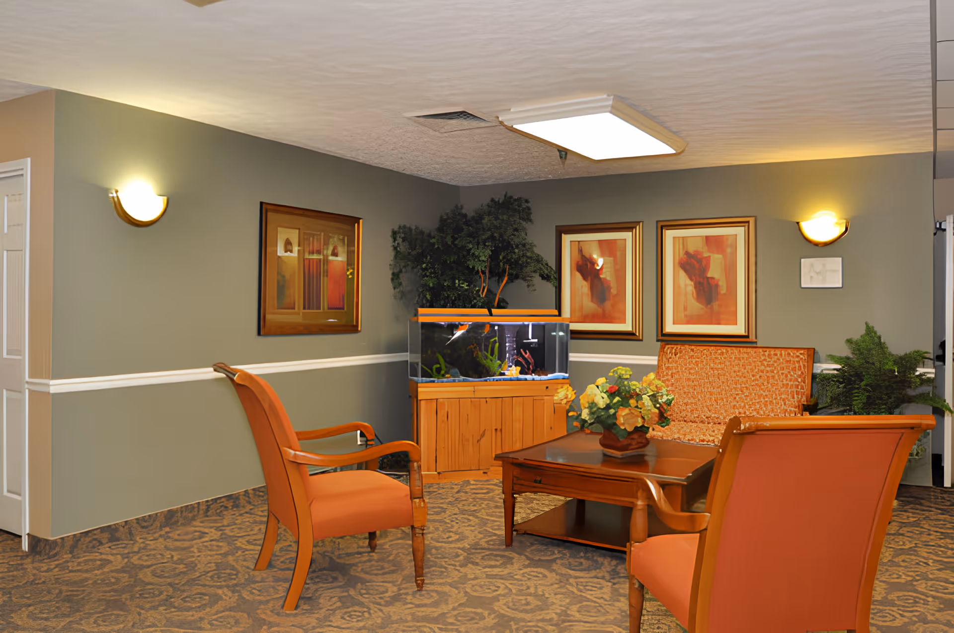 A cozy sitting area in a senior living facility with two orange armchairs and a patterned loveseat around a wooden coffee table with a flower arrangement. There is a large fish tank on a wooden stand against the wall, flanked by green plants. The walls are painted gray with white trim and decorated with framed artwork. The carpet has a floral pattern, and the room is softly lit by wall sconces and a ceiling light.