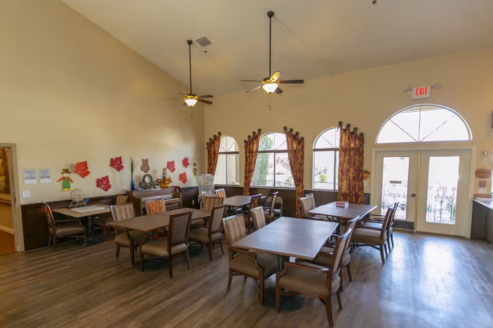 A dining room with several tables and chairs arranged neatly. The room has large arched windows with patterned curtains, ceiling fans with lights, and fall-themed decorations on the walls. There is a double door with glass panels leading outside.