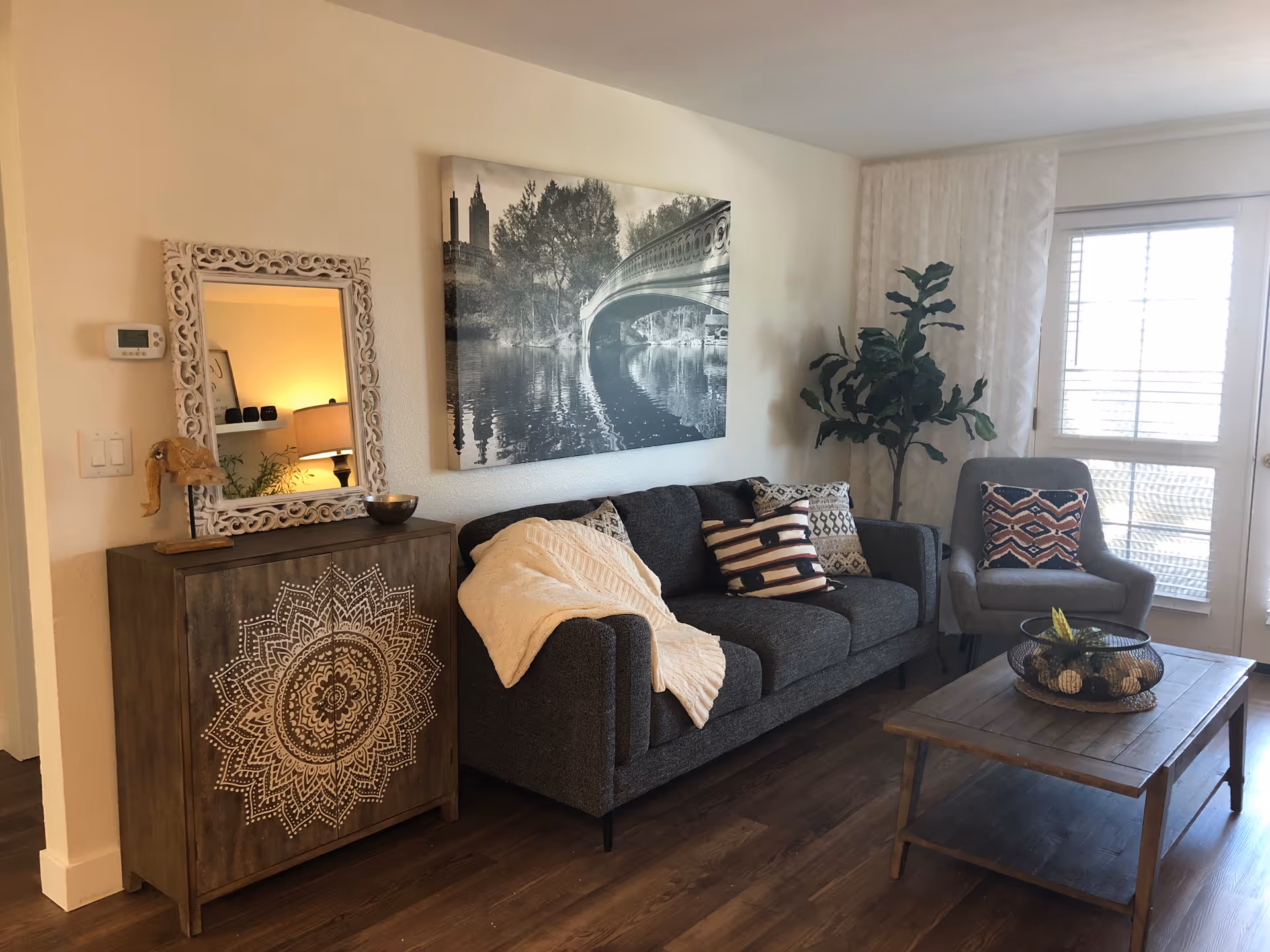 A cozy living room with a dark gray sofa adorned with patterned pillows and a cream-colored throw blanket. Next to the sofa is a wooden cabinet with an intricate white mandala design, topped with a decorative mirror and a small elephant figurine. A wooden coffee table with a decorative centerpiece sits in front of the sofa. A gray armchair with a patterned pillow is positioned near a window with white curtains and blinds. The walls are light-colored, and a large black and white photograph of a bridge over water hangs above the sofa.