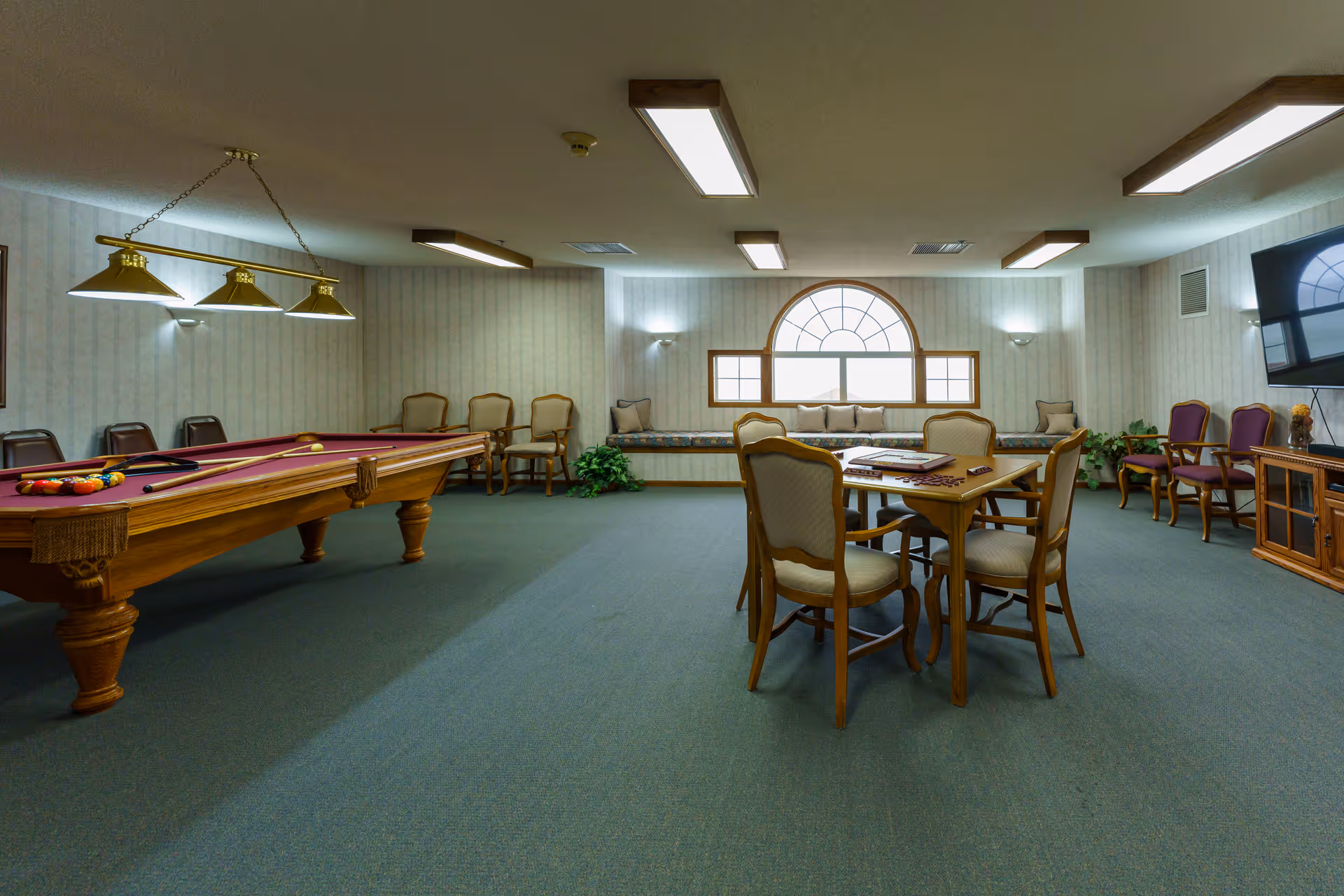 A senior living community game room with a pool table, card table and chairs, a TV, and a window seat under an arched window.