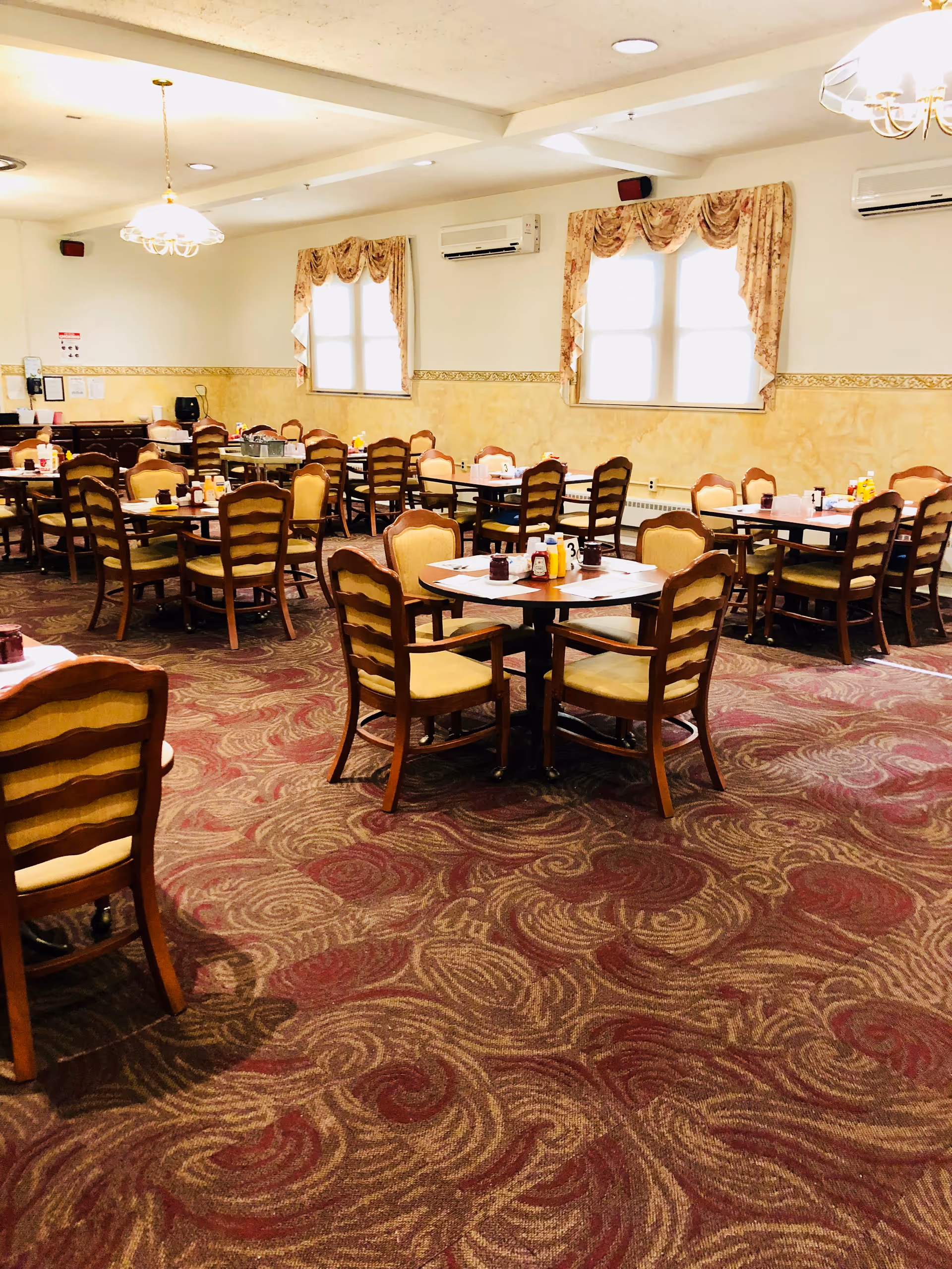 A dining room with multiple wooden tables and chairs arranged neatly. Each table has condiments and table numbers. The room has patterned carpet flooring, cream-colored walls with a decorative border, and windows with floral curtains. Ceiling lights provide illumination.