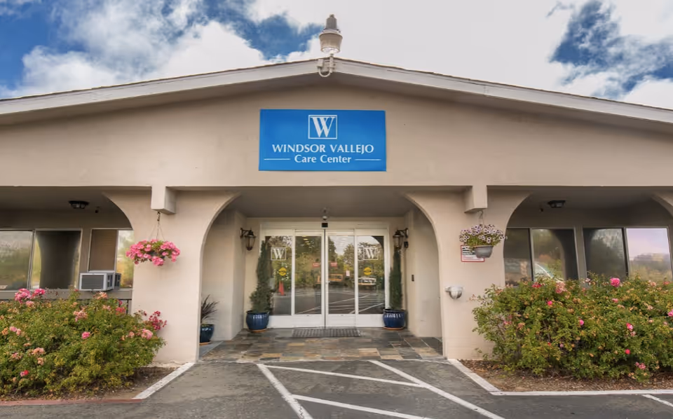 Entrance of Windsor Vallejo Care Center with a blue sign above double glass doors, potted plants, and flowering bushes.