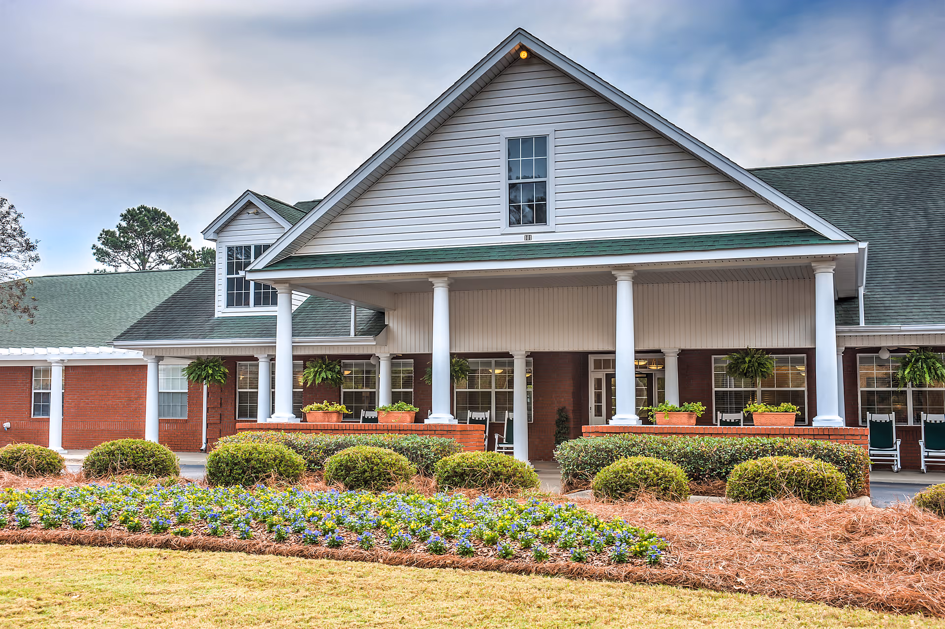Front exterior view of The Terrace at Grove Park building with a covered porch supported by white columns, green roof, brick walls, and landscaped garden with bushes and flowers in the foreground.
