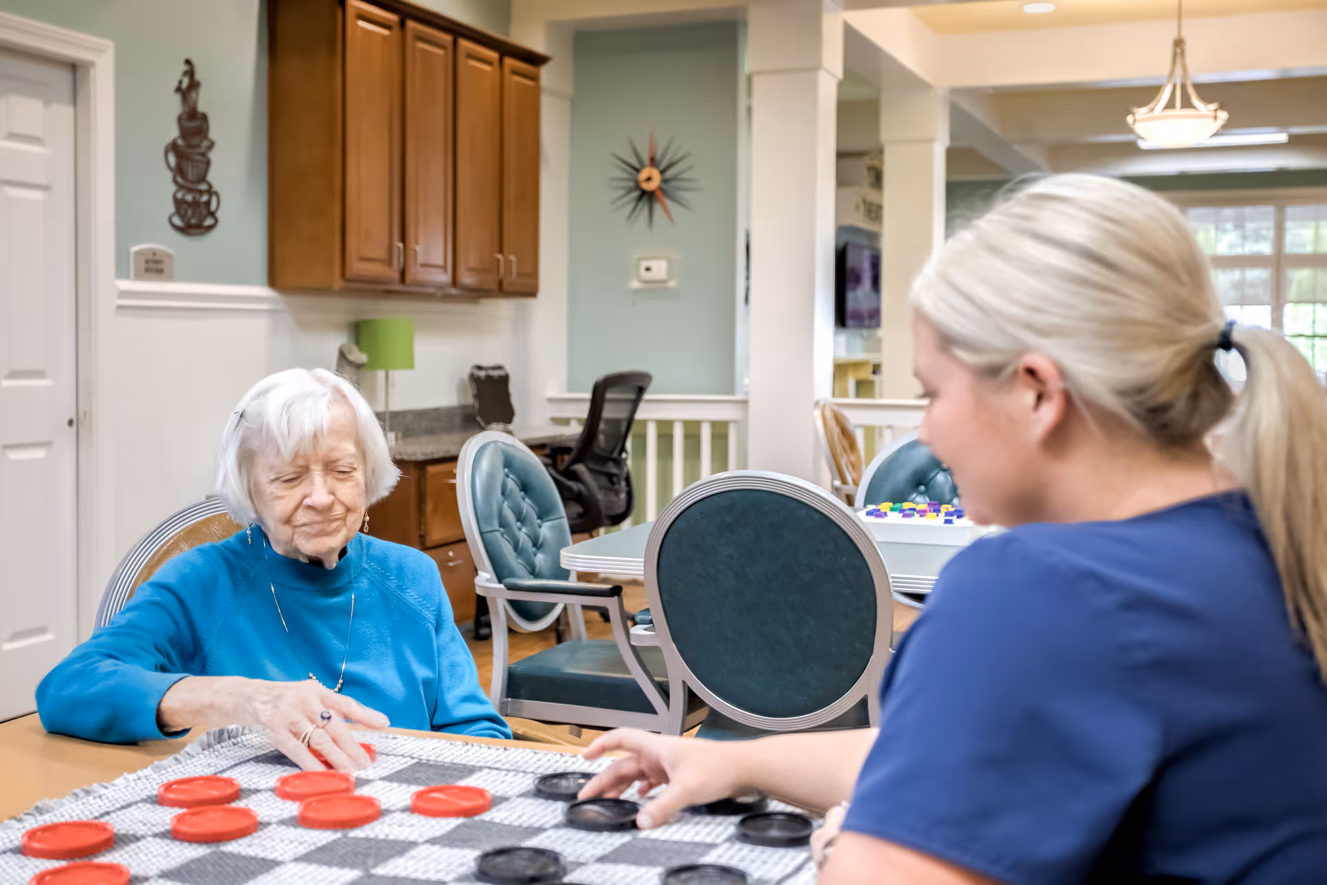 An elderly woman and a younger woman with blonde hair tied back are playing checkers at a table in a well-lit room with wooden cabinets, green cushioned chairs, and a decorative clock on the wall.