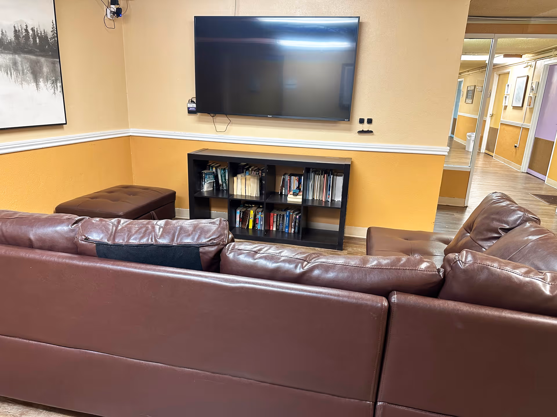 Seating area with brown leather sofas facing a wall-mounted TV above a small bookshelf in a common room.