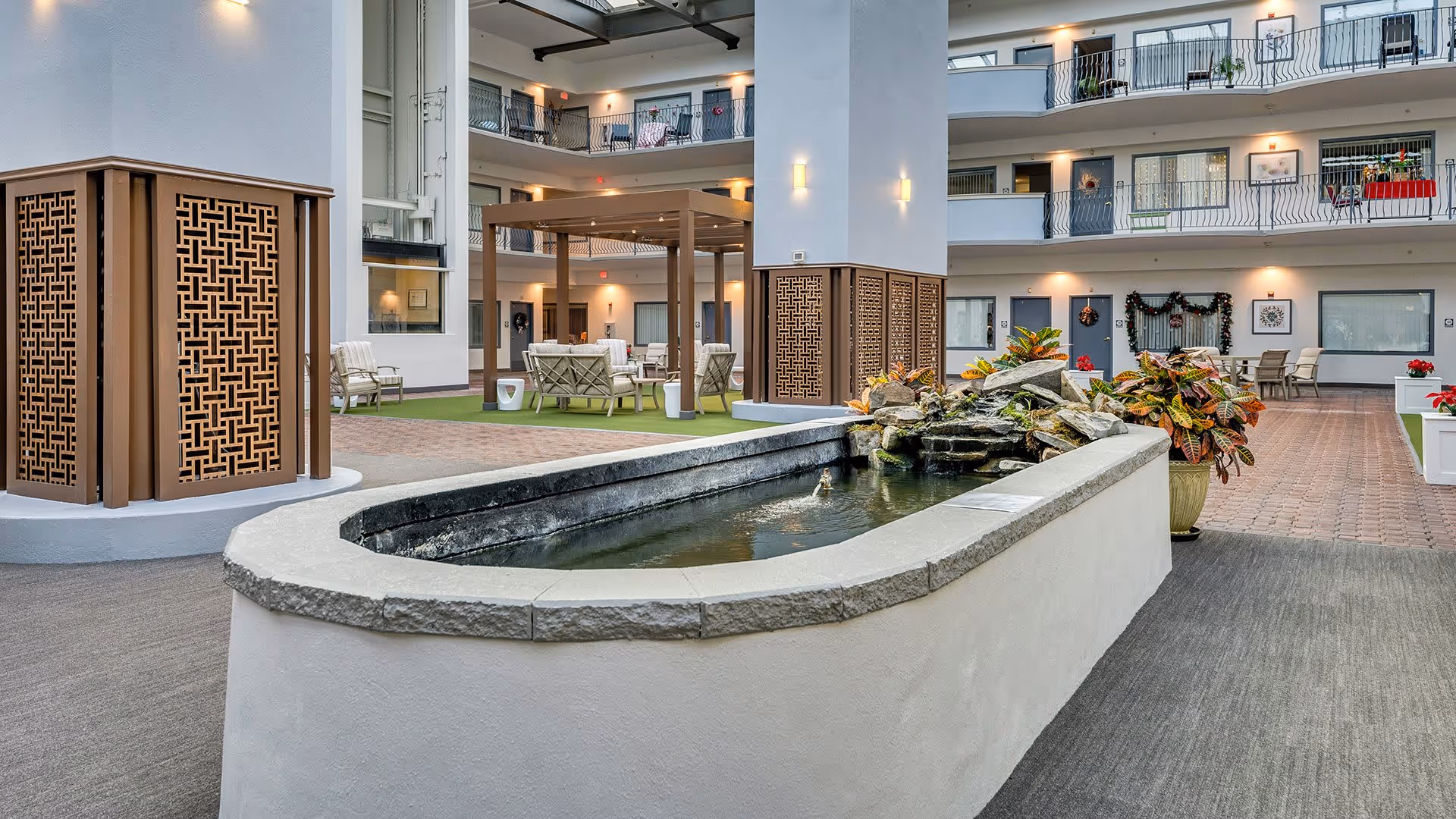 Indoor courtyard area of a senior living facility with a central water feature surrounded by plants. The space includes seating areas with chairs and tables under a pergola, and multiple floors with balconies overlooking the courtyard. The area is well-lit with wall sconces and decorated with holiday wreaths.