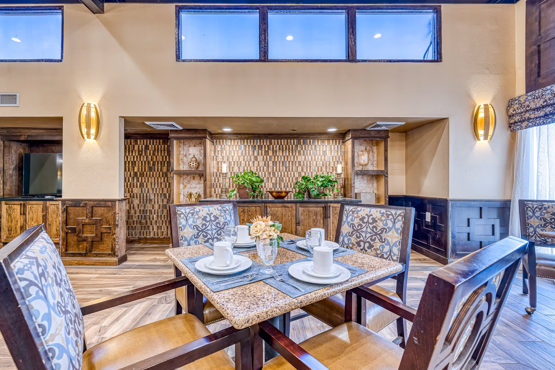 A well-lit dining area with a square table set for four, featuring patterned cushioned chairs, white cups, plates, and glassware. The background includes a decorative wooden cabinet with plants and ornamental items, warm wall sconces, and high windows letting in natural light.