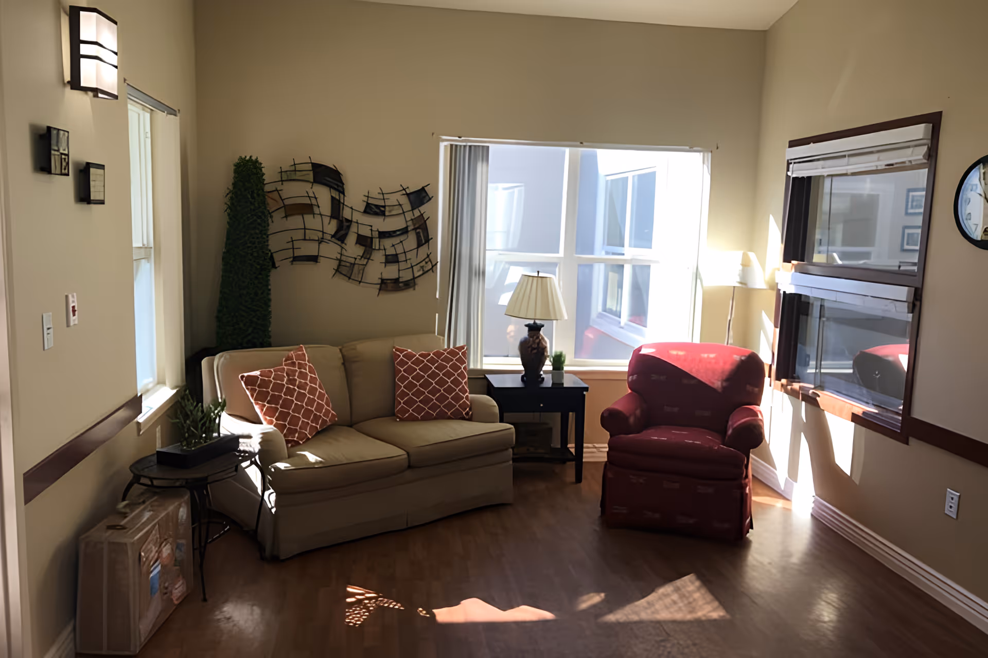 Sunlit living room with a beige sofa with patterned pillows, a red armchair, side tables, lamp and wall decor.