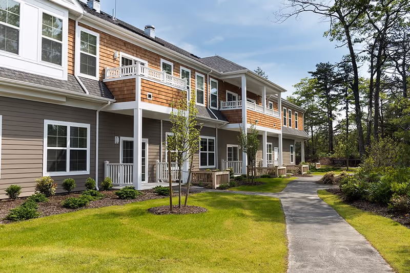 Exterior view of a two-story residential building with balconies and porches, surrounded by a well-maintained lawn, small trees, shrubs, and a paved walkway leading alongside the building. The sky is clear with some clouds, and there are tall trees in the background.