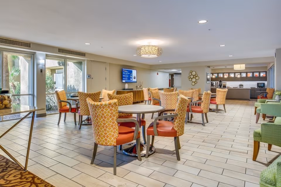 A spacious and well-lit dining area in a senior living facility featuring multiple round tables surrounded by orange and patterned chairs. The room has large windows allowing natural light, a modern ceiling light fixture, and a kitchenette area in the background with cabinets and appliances. Green armchairs are also visible on the right side.