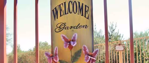 Wooden welcome sign reading Garden with butterflies at a gated outdoor entrance.