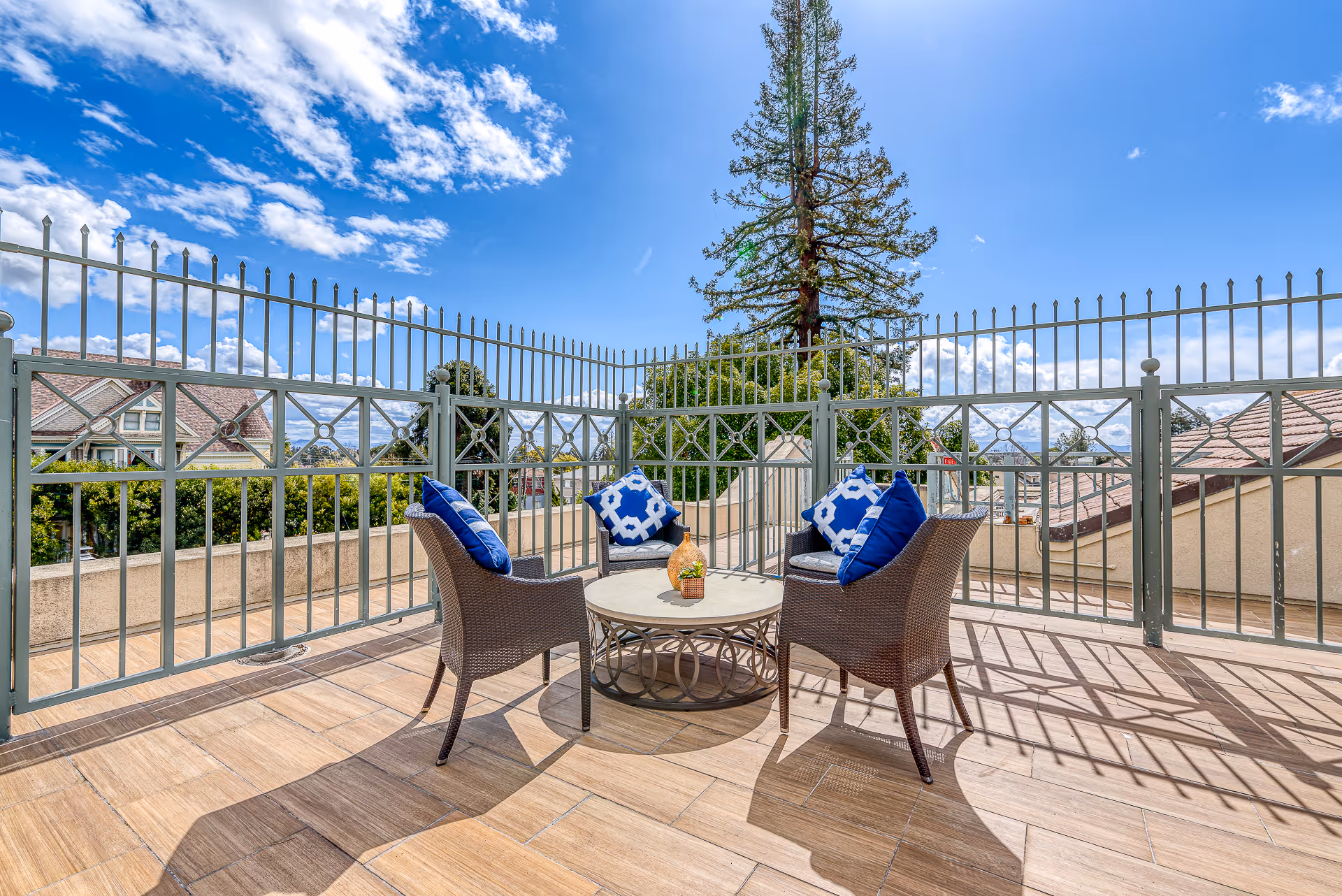 Rooftop patio with three wicker chairs around a round table and blue patterned cushions, enclosed by decorative metal fencing under a bright blue sky.