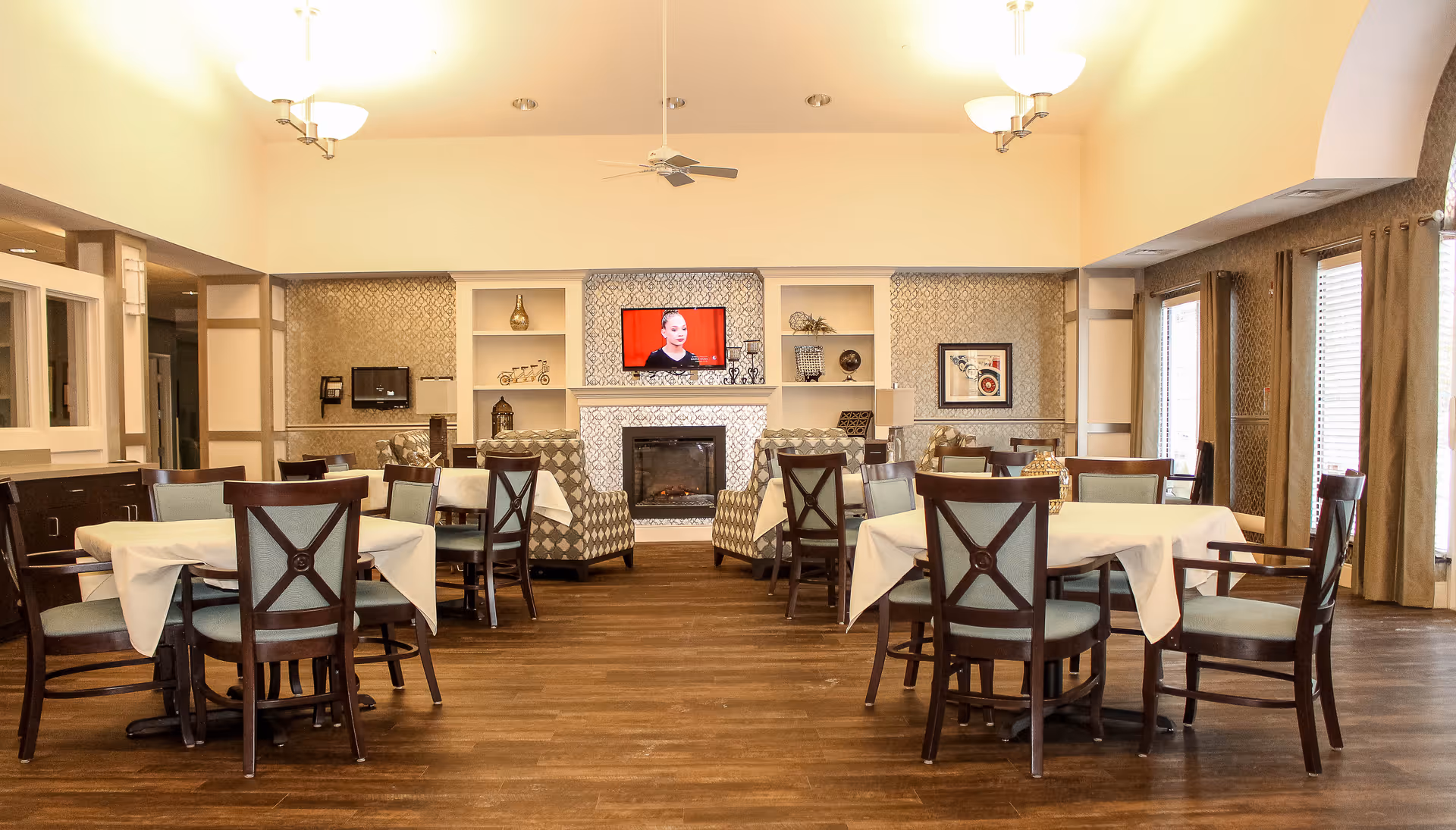 A well-lit dining room with several tables covered with white tablecloths and surrounded by wooden chairs with light blue cushions. The room features a fireplace with a TV mounted above it, flanked by built-in shelves with decorative items. Large windows with curtains allow natural light to enter, and the floor is made of wood.