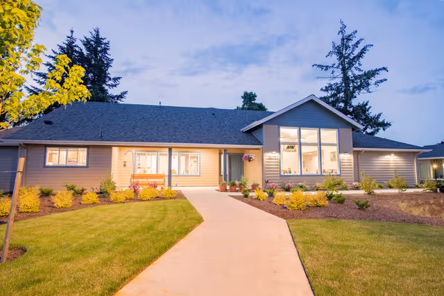 Exterior view of a single-story residential building with a well-maintained lawn and landscaped garden beds along a concrete walkway leading to the entrance. The building has large windows and is illuminated with warm lighting during dusk.