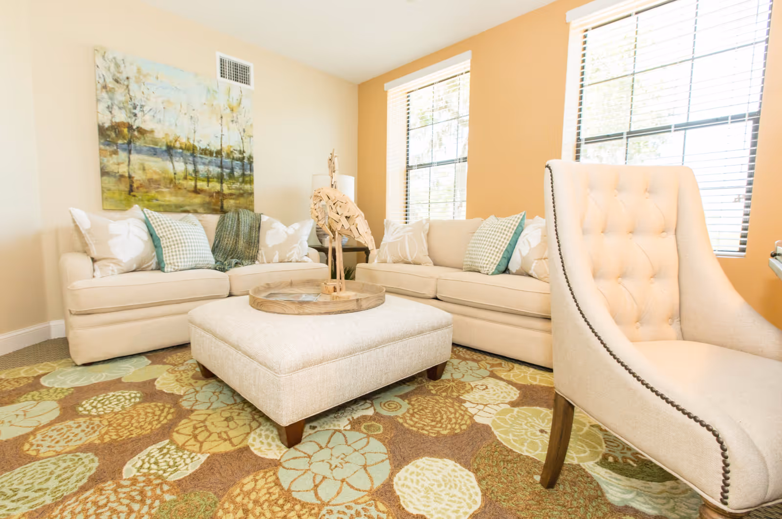 Bright, furnished living room with beige sofas, a tufted armchair, ottoman coffee table and a patterned rug.