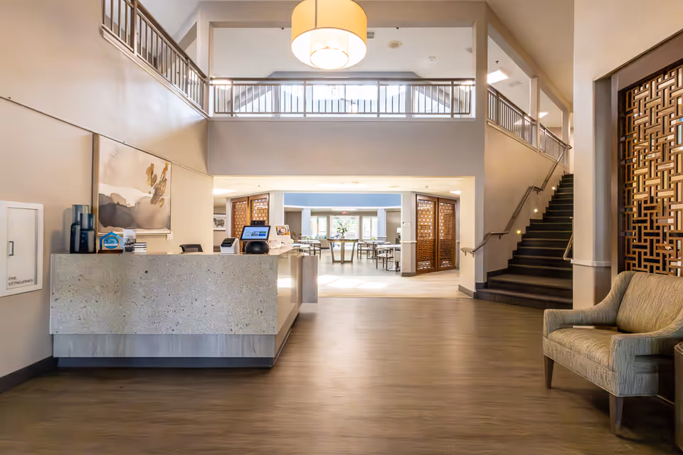 Reception area of Asher Point Independent Living of Arlington with a modern front desk, a large hanging light fixture, a staircase to the right, and a seating area with a chair. The space is well-lit with natural light coming from the dining area visible in the background.