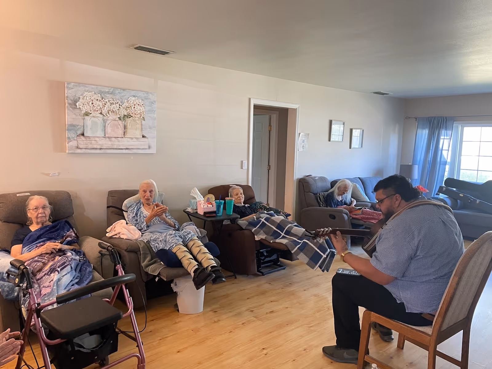 A group of elderly women sitting in recliners in a living room, some covered with blankets. A man is sitting on a chair playing a guitar for them. The room has light-colored walls, wooden flooring, a painting of flowers on the wall, and a window with blue curtains letting in natural light.