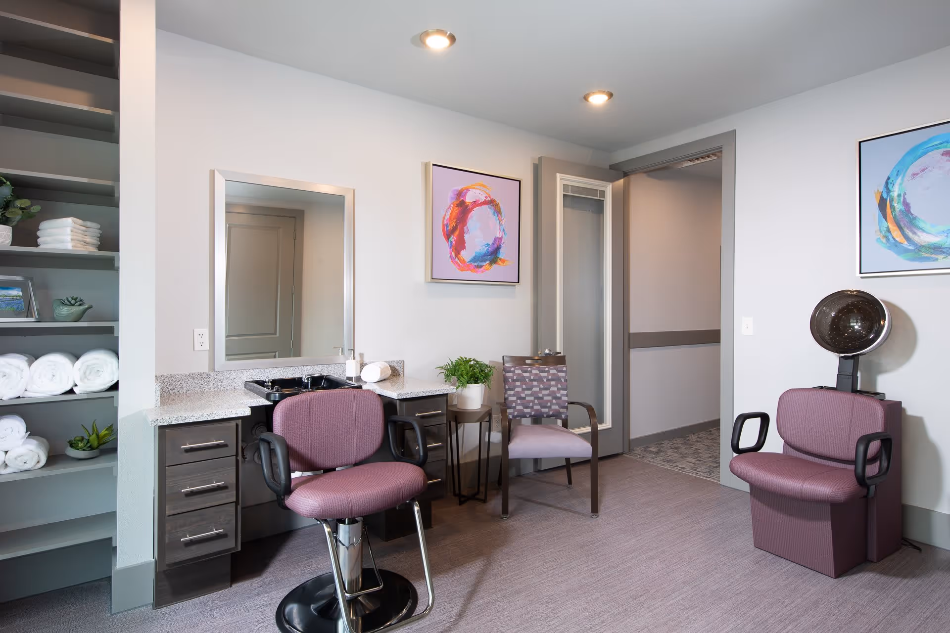 Interior view of a salon area in a senior living facility with a purple salon chair in front of a mirror and counter, a patterned chair next to a small table with a potted plant, shelves with rolled white towels and decorative items, and a hair dryer chair with a hooded dryer. The walls are light gray with colorful abstract paintings.