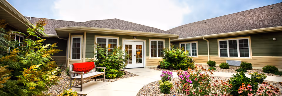 Outdoor courtyard area of a senior living facility with a curved concrete walkway, garden beds filled with various plants and flowers, a wooden bench with a red cushion, and a building with green and beige siding and multiple windows under a cloudy sky.