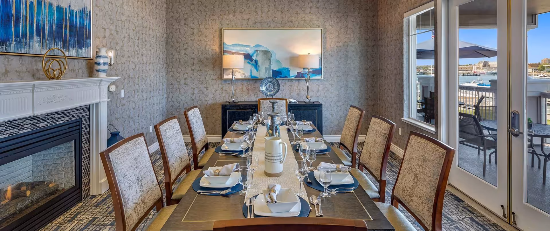 A formal dining room with a long table set for eight people. The table is arranged with white plates, napkins, silverware, and glassware. The room features patterned wallpaper, a white fireplace with decorative items on the mantel, a sideboard with two lamps and a decorative plate, and large windows and glass doors that open to an outdoor patio with a view of a waterfront and boats.