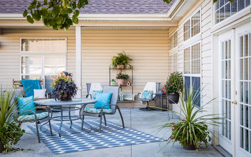 Outdoor patio area with a round glass table surrounded by four cushioned chairs, some with blue and patterned pillows. The patio is decorated with potted plants and flowers, a patterned outdoor rug, and a small shelving unit with more plants and decorative items against the beige siding of the building. There are large windows and glass doors on the right side.