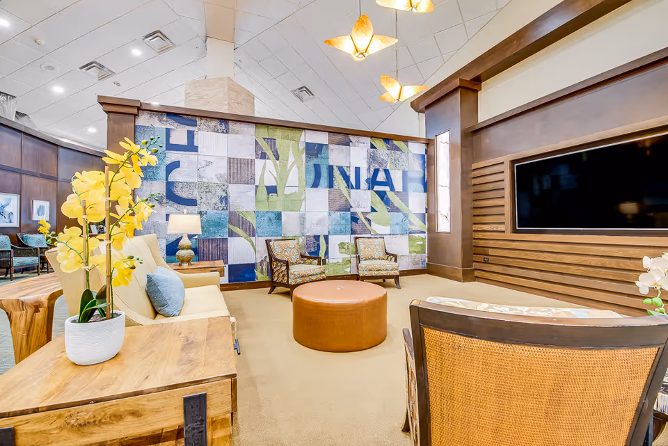 A bright and spacious lounge area in a senior living facility with comfortable seating including armchairs and a sofa, a large wall-mounted flat screen TV, decorative yellow orchids on wooden tables, and a colorful tiled wall with abstract patterns. The room features warm lighting and wood paneling.