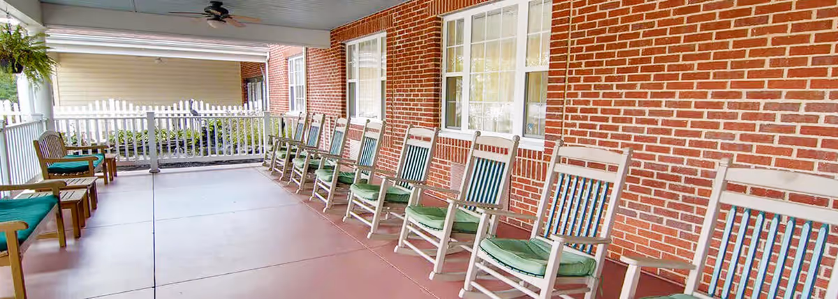 Covered outdoor patio area at Milford Place with a row of white rocking chairs with green cushions lined up against a red brick wall, a ceiling fan above, and a white picket fence enclosing the space.
