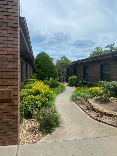 A concrete pathway flanked by well-maintained green shrubs and small trees, running between two single-story brick buildings under a partly cloudy sky.